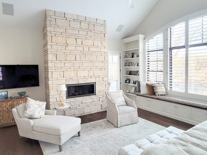 a cream colored natural stone fireplace laid in a coursed ashlar pattern in an all white bedroom with large, bright windows on one wall